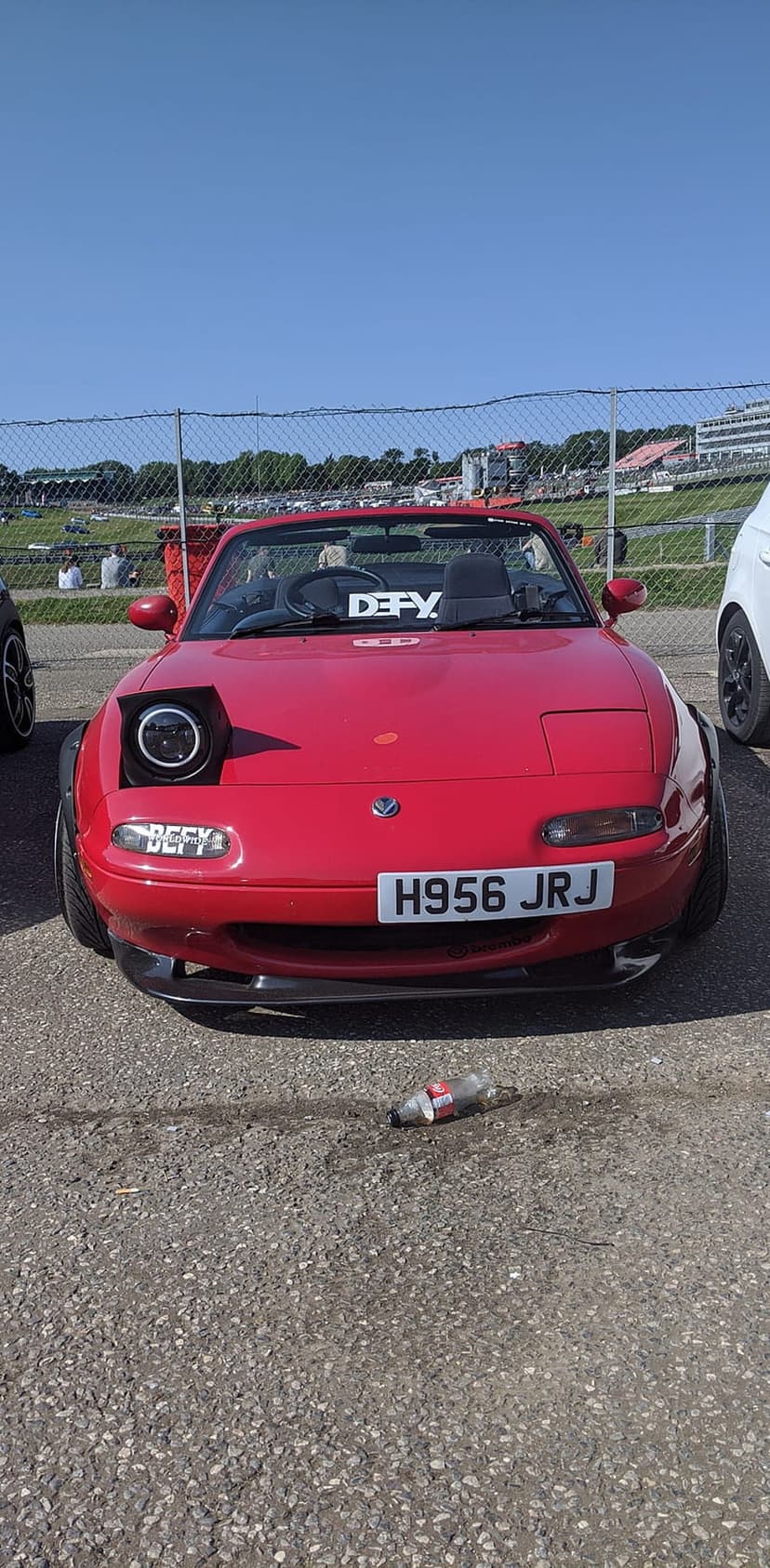 Red Mazda Miata with one pop-up headlight open and widebody fenders parked on asphalt at a sunny car event.