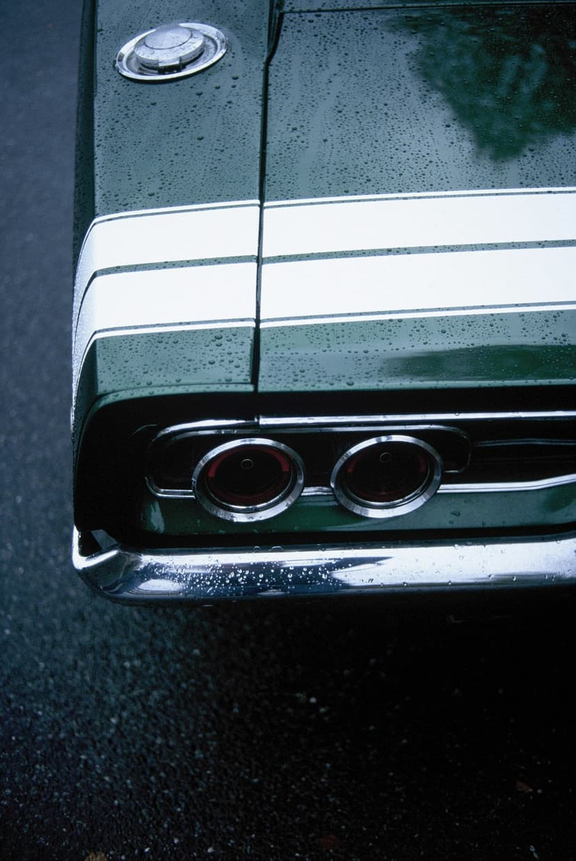 Rear of a wet vintage green car with white racing stripes, round taillights, and chrome bumper in the rain.