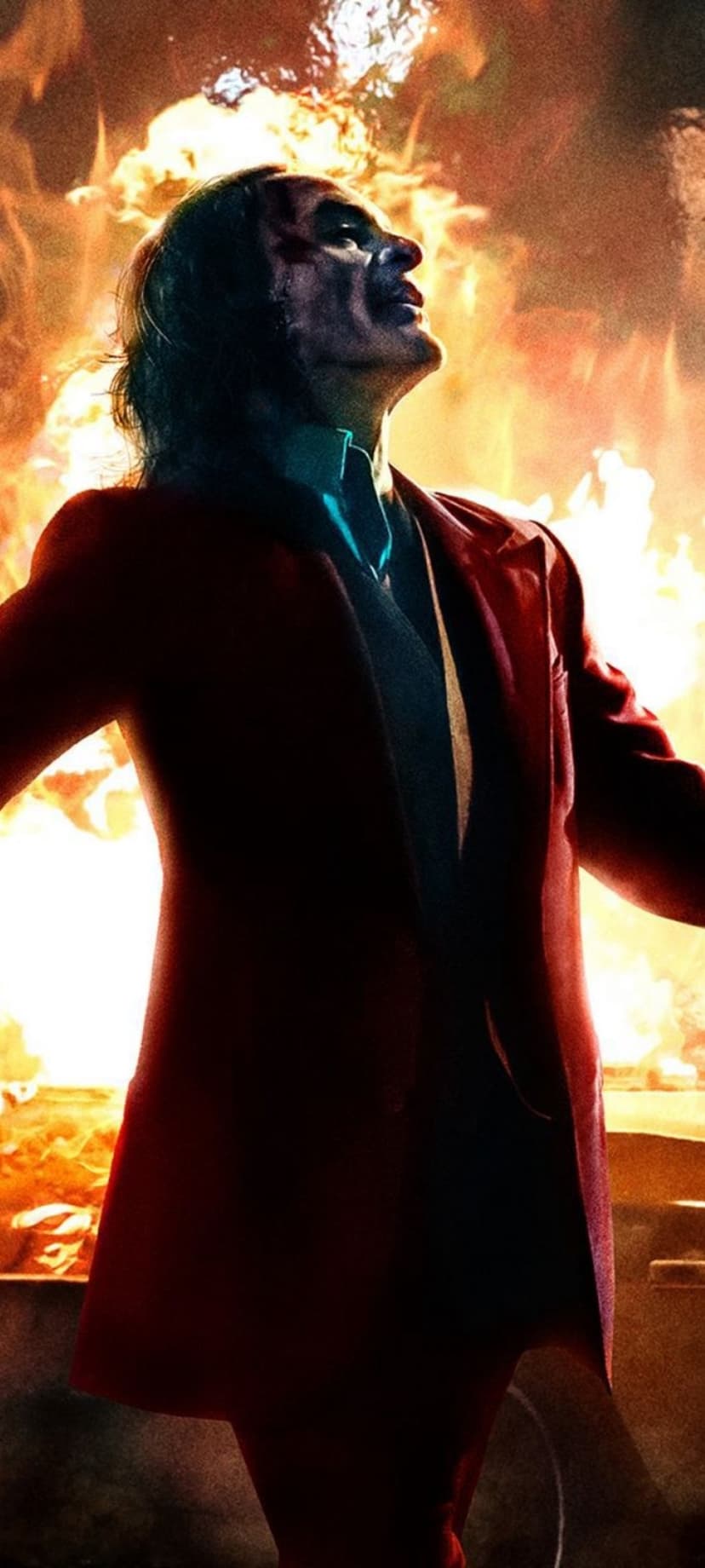 Arthur Fleck in a red suit and clown makeup looks upward as a giant fire explosion erupts behind him.
