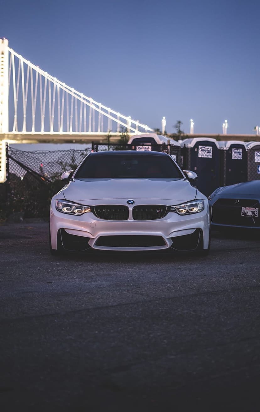 White BMW M4 sports car parked in front of a golden-lit suspension bridge under a dark blue evening sky.