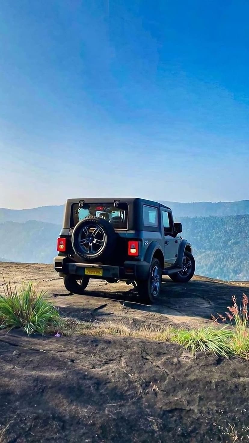 Mahindra Thar SUV parked on a rocky cliff edge overlooking green mountain ridges under a bright blue sky.