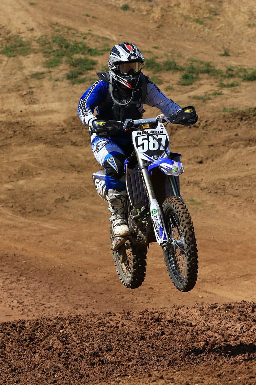 Motocross rider in blue and white gear jumps a dirt bike over a dusty track mound under a bright sky.