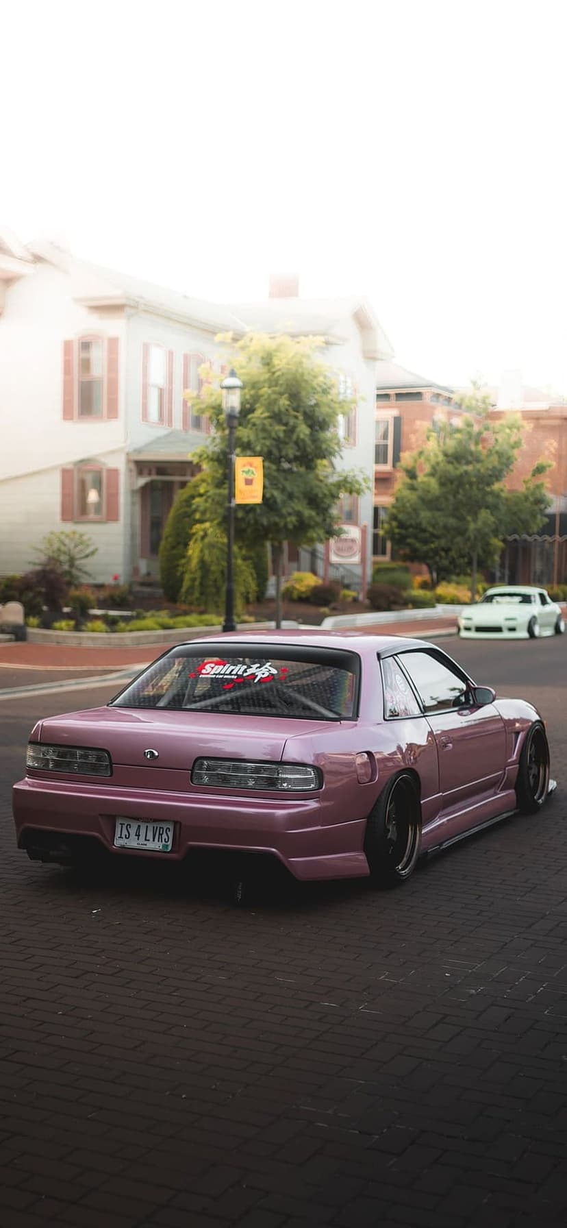 Pink Nissan 240SX S13 drifting on cobblestones past a parked green car and houses in afternoon light.