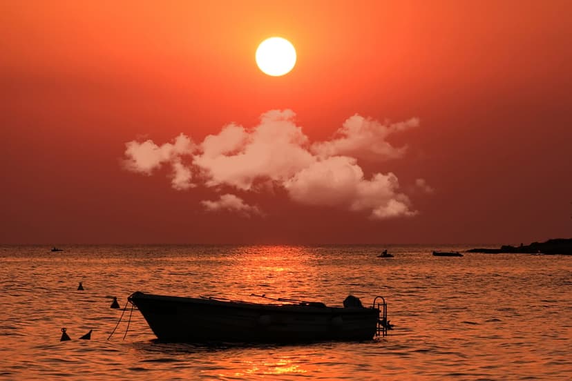 Lone boat silhouetted against a fiery orange and red sunset over calm ocean waters with wispy clouds.