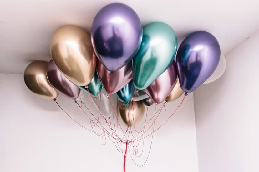 Iridescent purple, gold, and teal chrome balloons cluster against a white ceiling with thin red strings.
