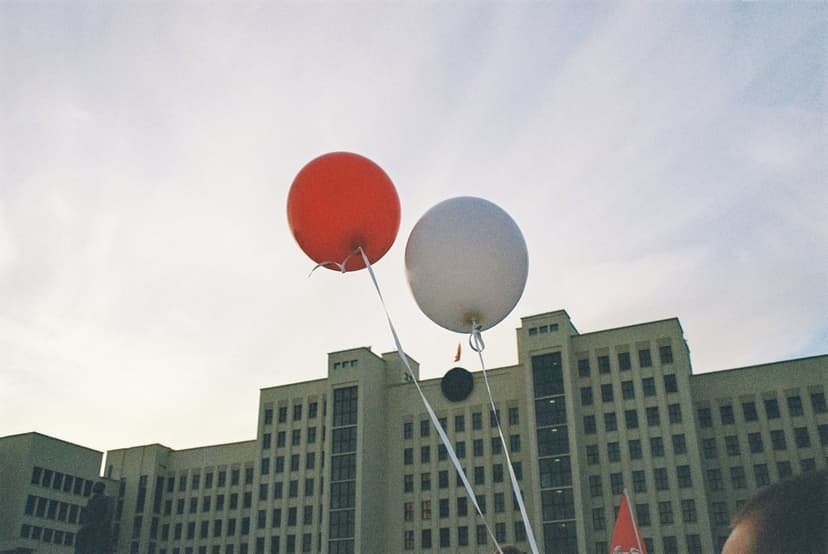 Red and white balloons with trailing ribbons drift in a cloudy sky above a large, multi-story stone building.