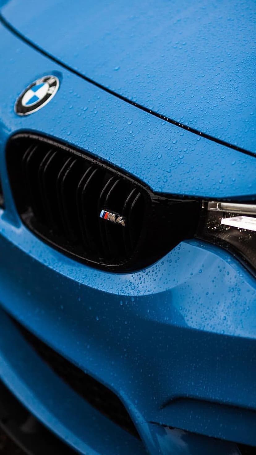 Blue BMW M4 front grille and emblem covered in water droplets with reflections on the metallic paint surface.