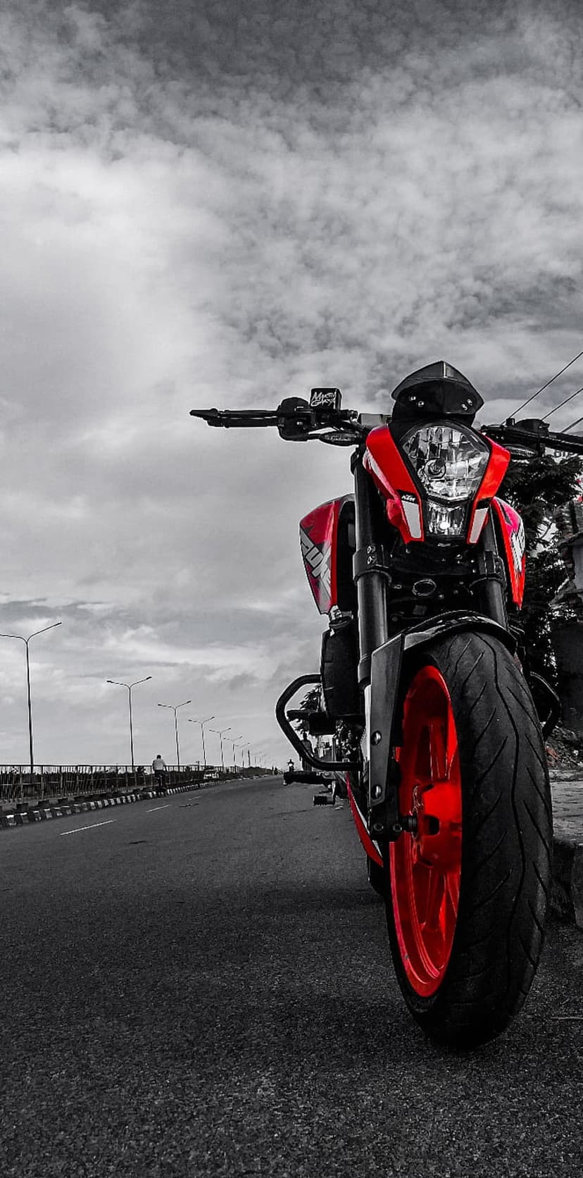 Red motorcycle parked on a grayscale asphalt road beneath a dramatic cloudy sky and distant streetlights.