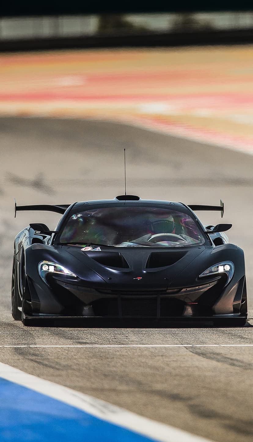 Black McLaren P1 GTR supercar with glowing headlights speeds across a blurred asphalt race track circuit.