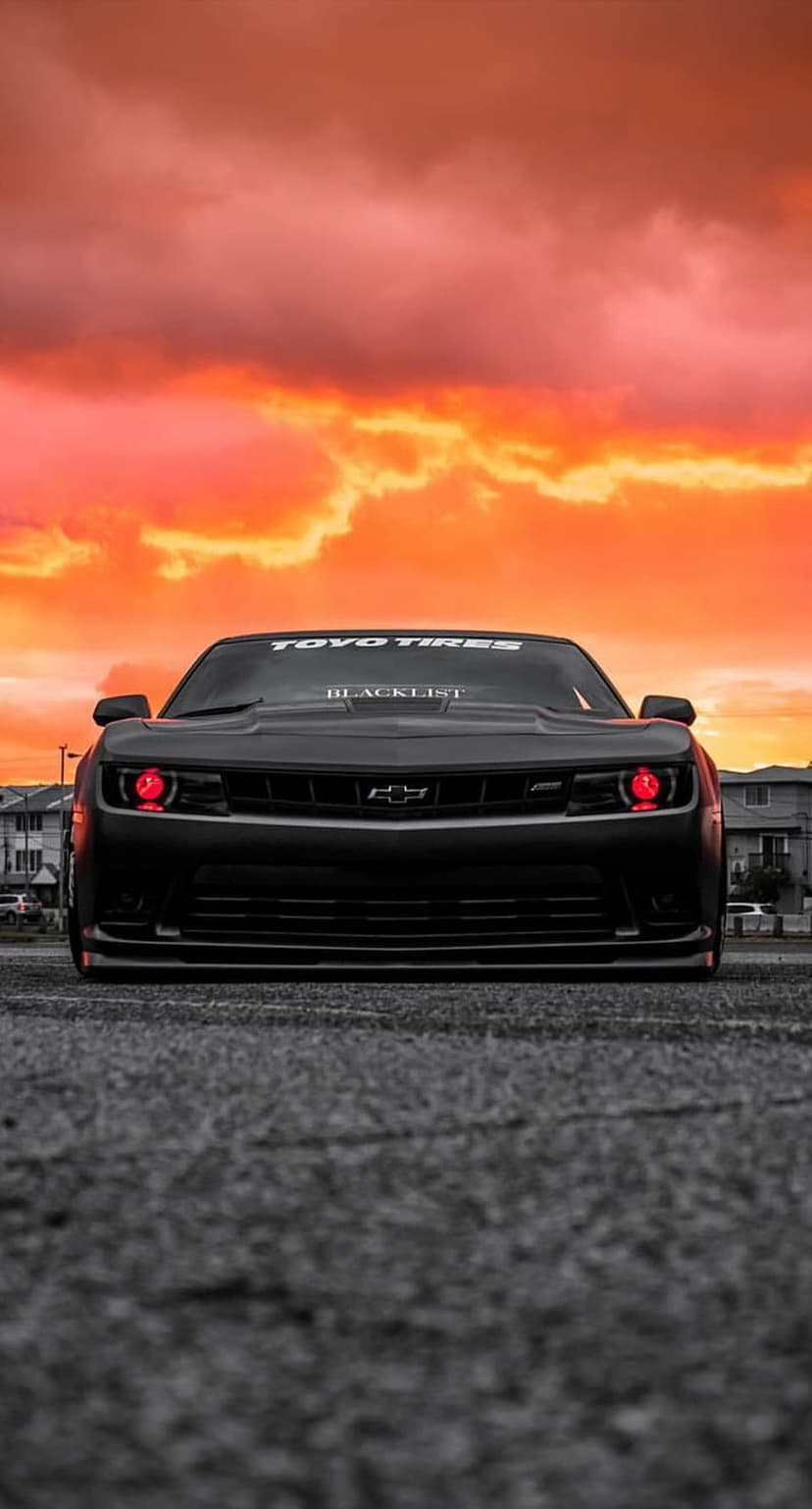 Black Chevrolet Camaro with glowing red headlights on asphalt under an orange and pink sunset sky at twilight.
