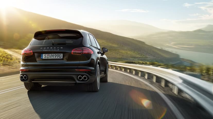Black Porsche Cayenne Turbo S rear view driving on a winding mountain road with golden hour sun flare and valley.