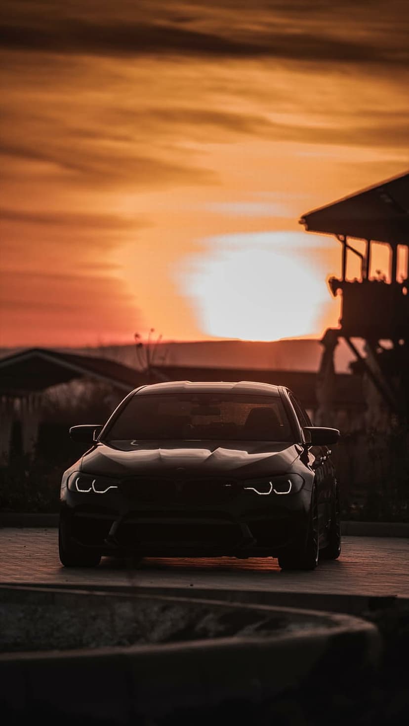 Black luxury car with glowing headlights parked on asphalt against a vibrant orange and yellow sunset sky.