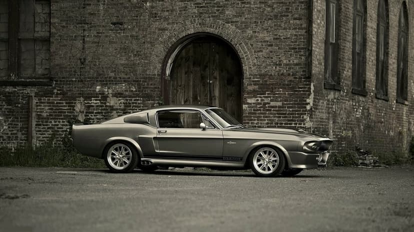Silver 1967 Shelby Mustang GT500 parked in front of a weathered brick wall and arched wooden industrial door.
