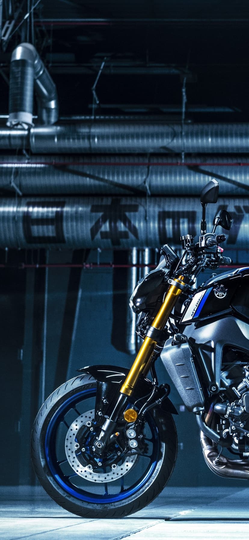Front of a black motorcycle with gold forks and blue rims in a dark industrial pipe-filled environment.