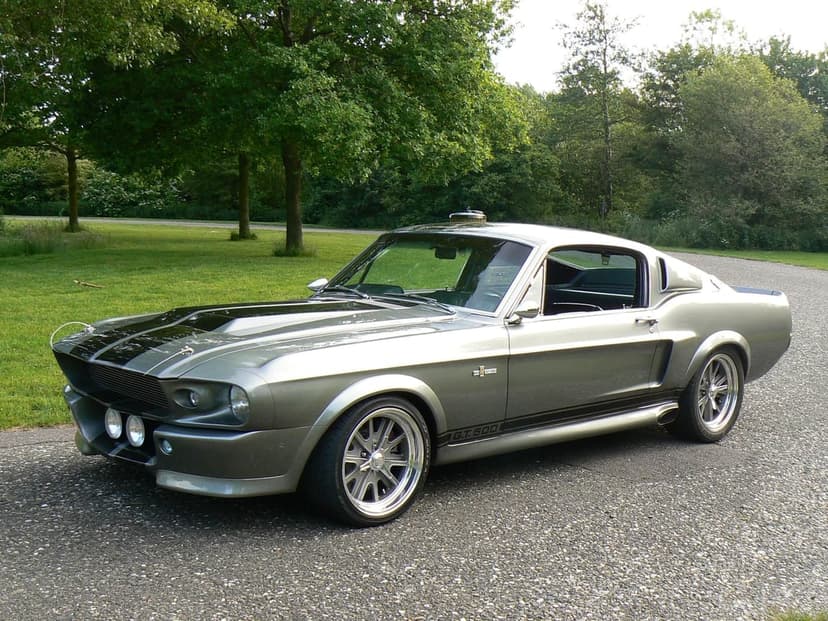 Metallic silver Shelby GT500 Eleanor with black racing stripes parked on a road surrounded by green park trees.
