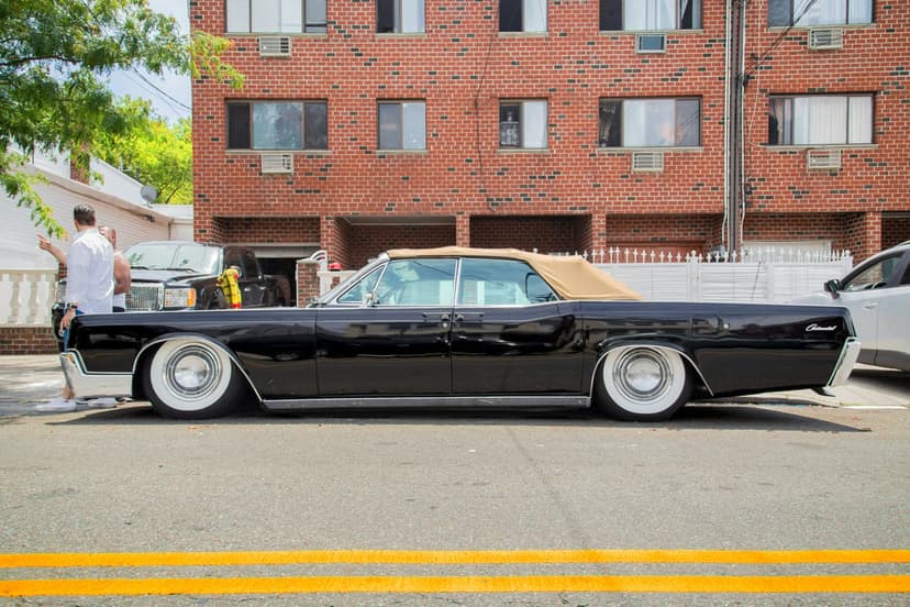 Black 1960s Lincoln Continental convertible with tan top and chrome trim parked beside a brick urban building.