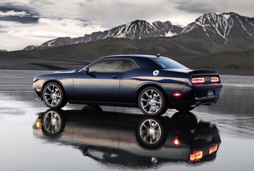 Dark blue Dodge Challenger SRT parked on wet asphalt with snowy mountain reflections under a moody overcast sky.