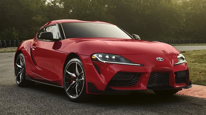 Red Toyota Supra GR sports car parked on an asphalt racetrack with green trees and a tire wall in the background.