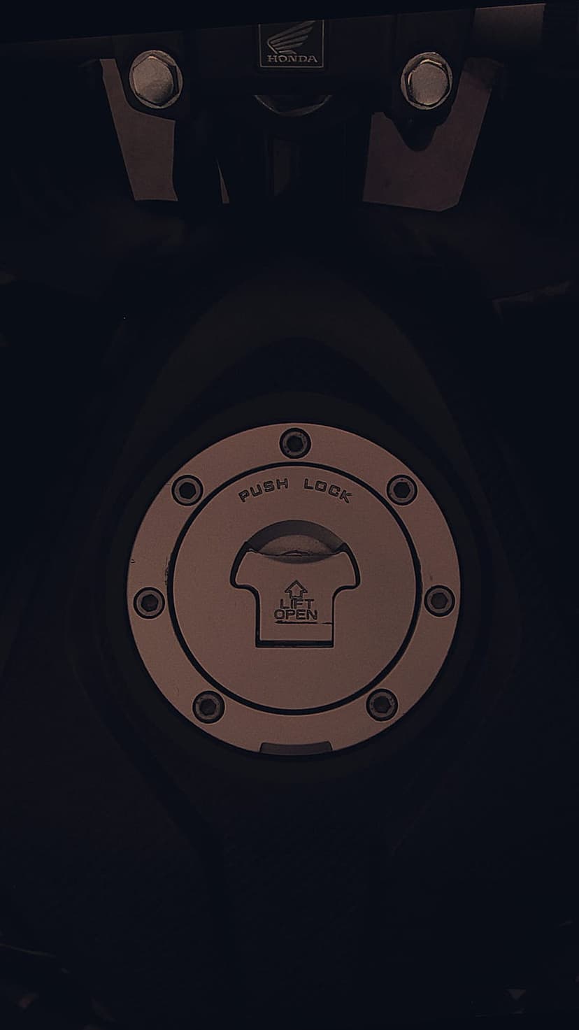 Honda fuel cap with circular push lock engraving and brushed metal texture on a vertical motorcycle gas tank.