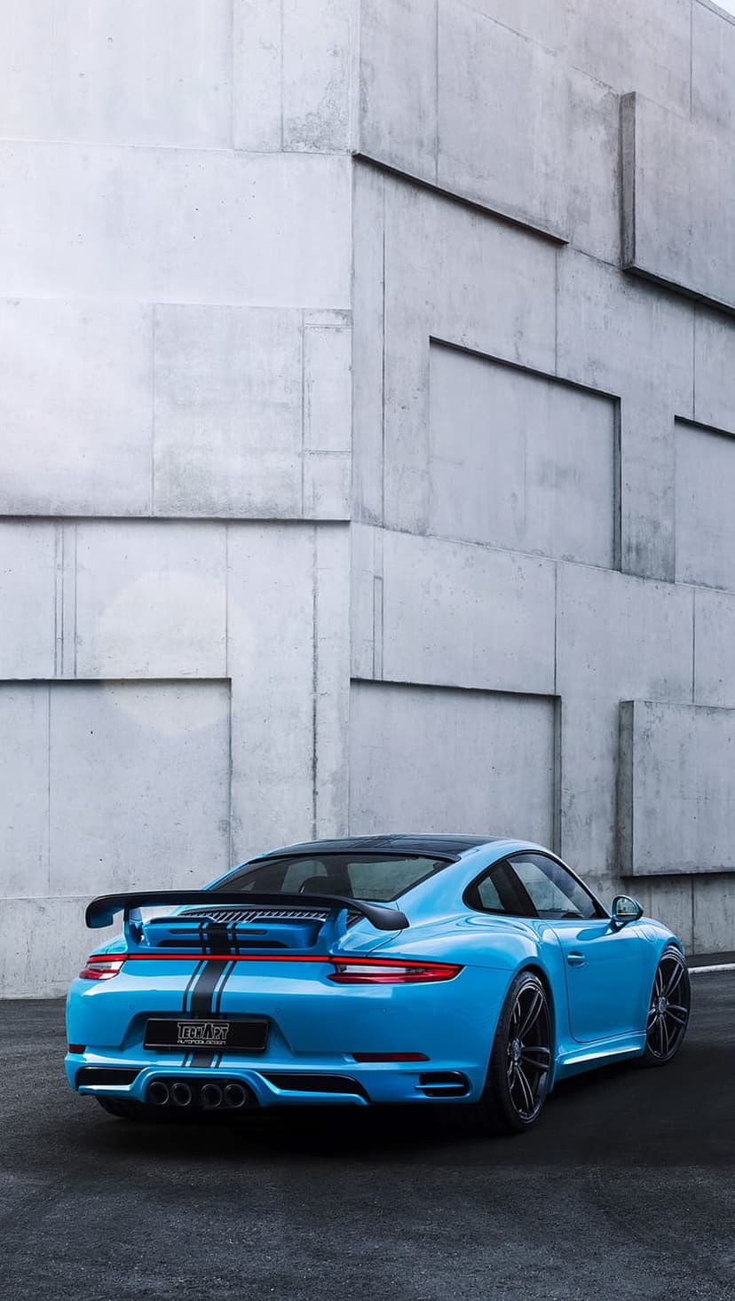 Blue Porsche 911 rear view with black racing stripes and a large wing parked in front of a concrete building.
