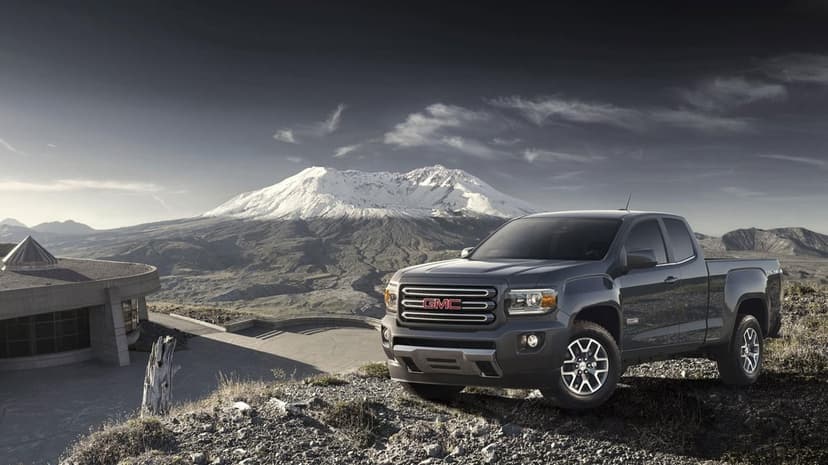 Dark grey GMC pickup truck on a rocky ridge with a massive snow-covered mountain peak under a cloudy sky behind it.