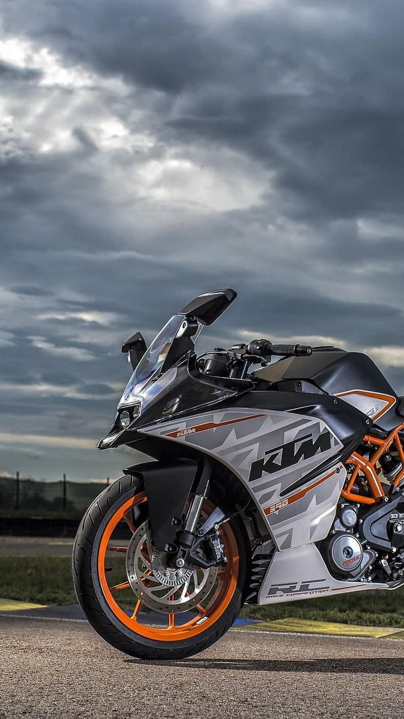 KTM RC 390 motorcycle with an orange wheel sits on asphalt under a dramatic, cloudy sky at a racing circuit.