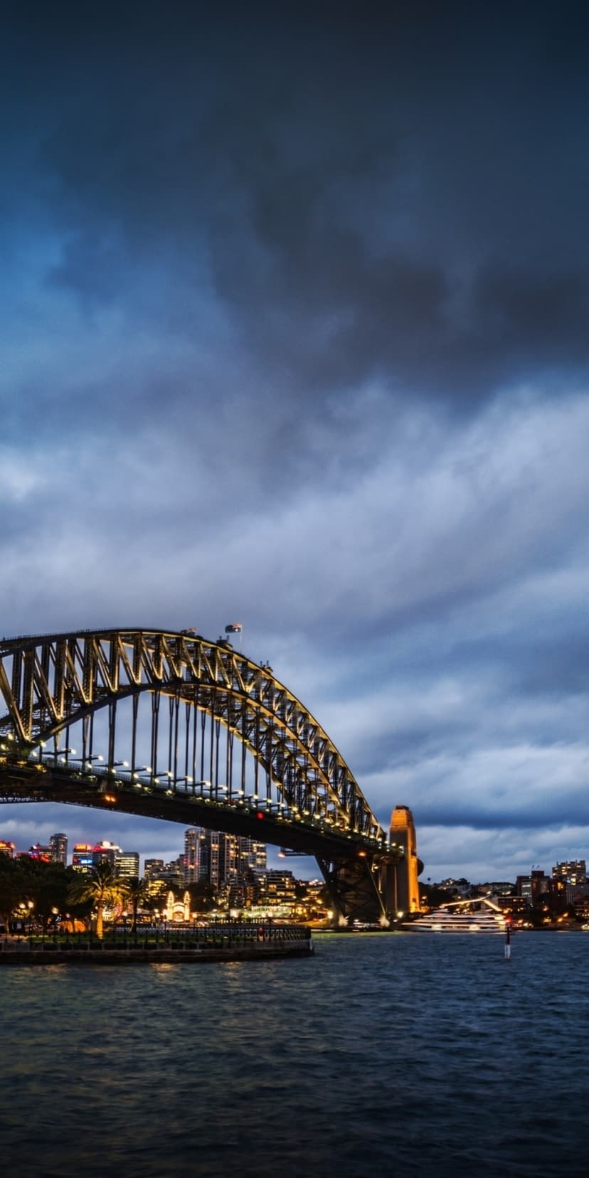 Sydney Harbour Bridge glows with amber lights against a dark blue twilight sky over reflective harbor waters.