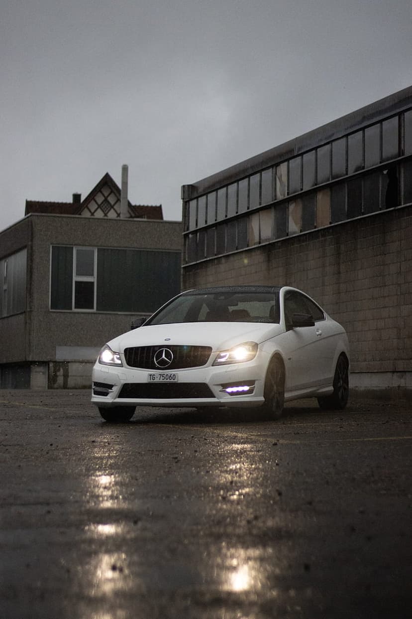 White Mercedes coupe with glowing headlights on a wet city street under a dark, overcast sky after a rainstorm.
