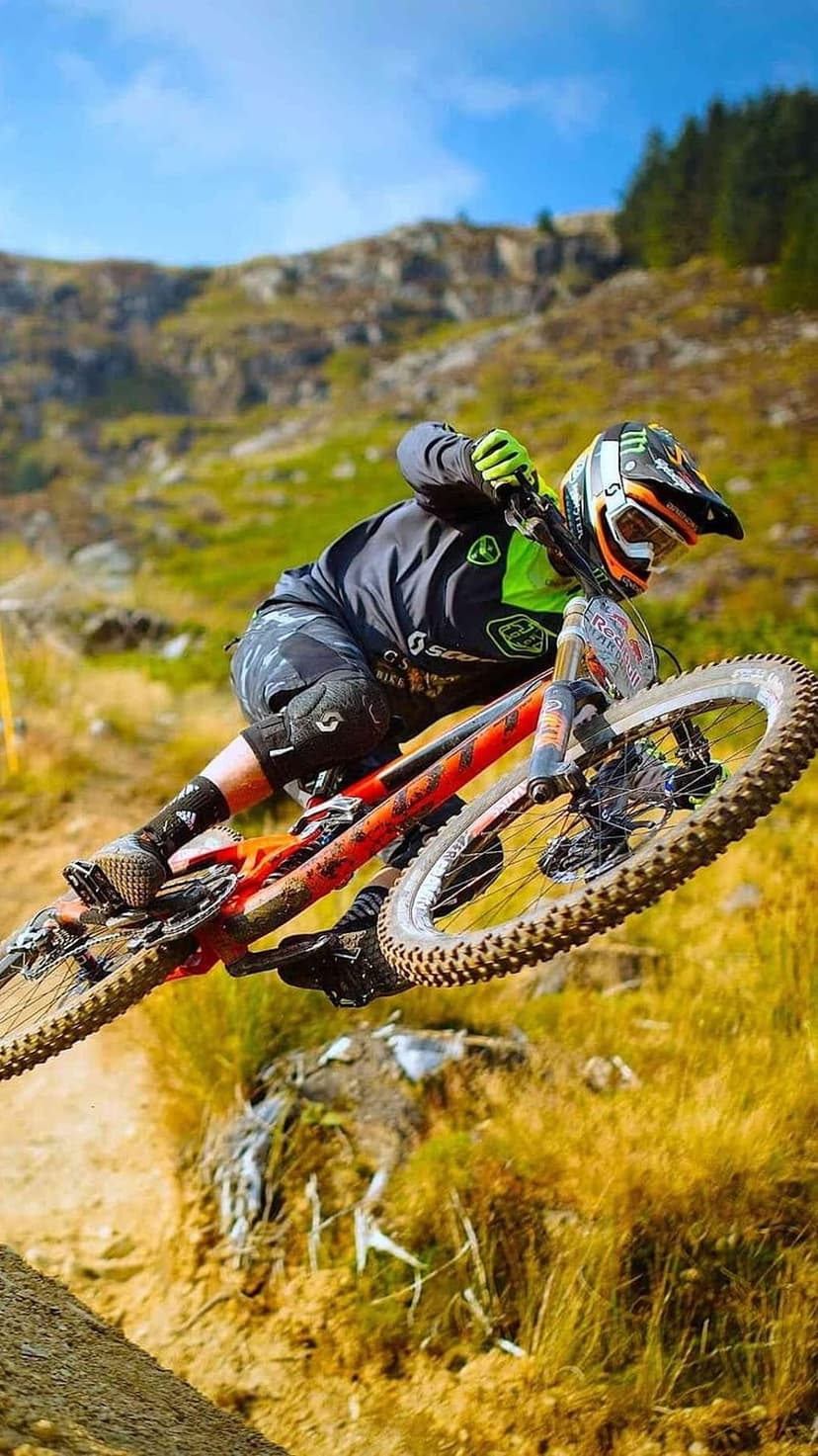 Mountain biker mid-air on a red downhill bike over grassy slopes under a blue sky with scattered white clouds.