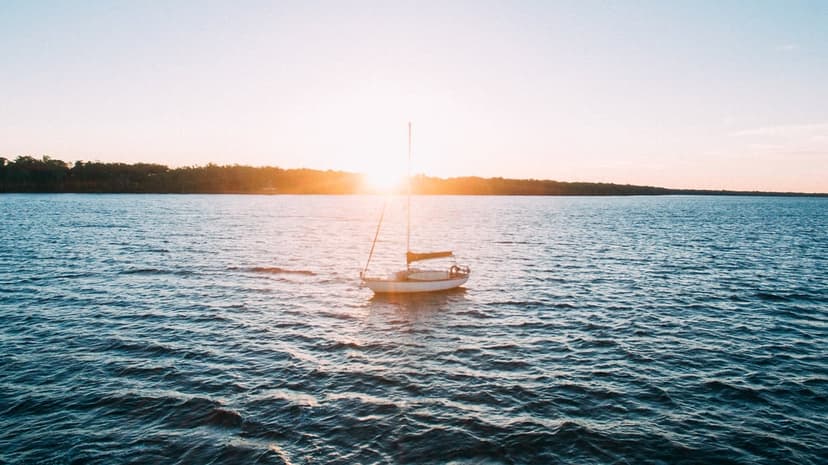 White sailboat drifting on a glassy lake under an orange sunset sky with vibrant golden water reflections.