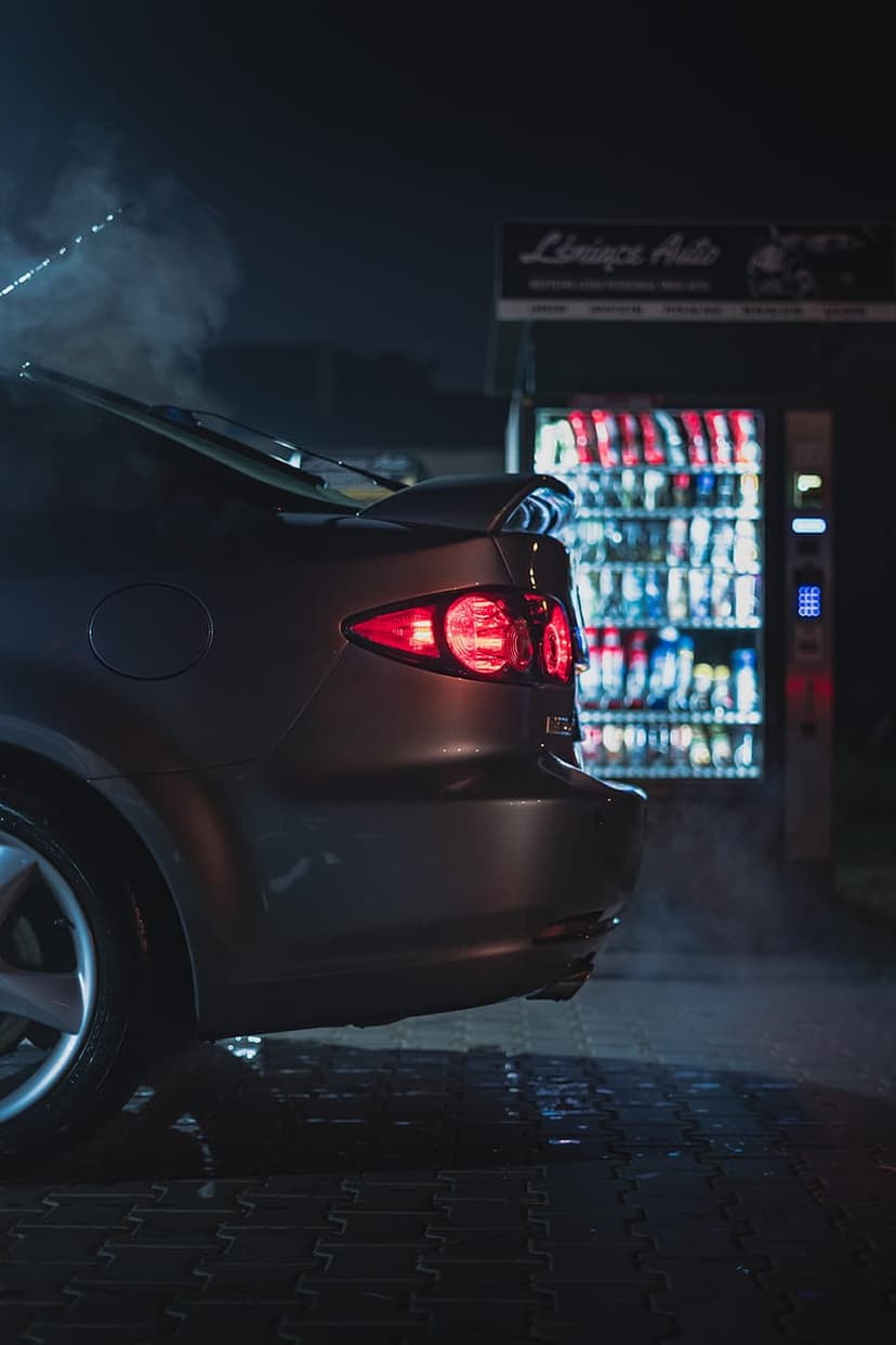 Black car idles next to a glowing vending machine at night with steam rising from the exhaust pipe on a city street.