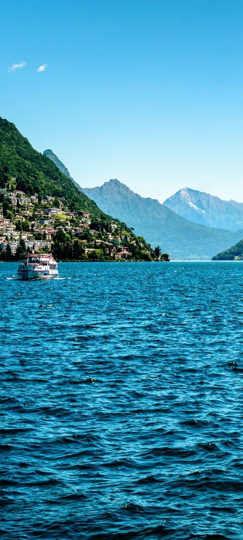 White pleasure boat sails past colorful houses on a steep mountainside overlooking the blue waters of Lake Como.