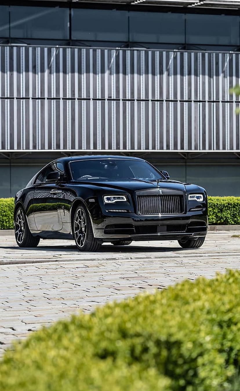 Black Rolls-Royce Wraith parked on a paved driveway in front of a modern building with silver vertical panels.