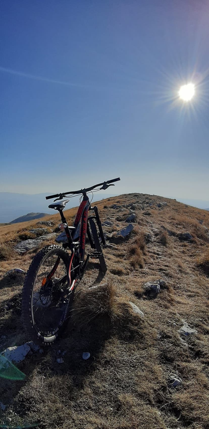 Mountain bike parked on a grassy ridge under a bright sun with hazy distant peaks and a clear blue sky.