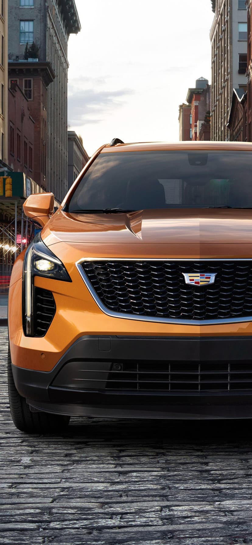 Metallic bronze Cadillac SUV front profile on a cobblestone street with urban buildings in the background.