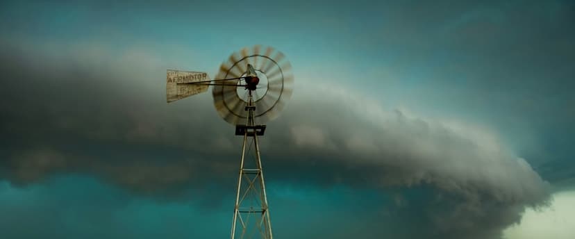 Windmill with blurred blades stands against swirling dark teal and gray storm clouds in a moody rural field.