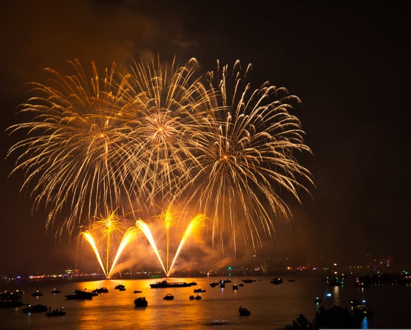 Golden fireworks bursts exploding in a dark night sky with reflections shimmering on the water and distant boats.