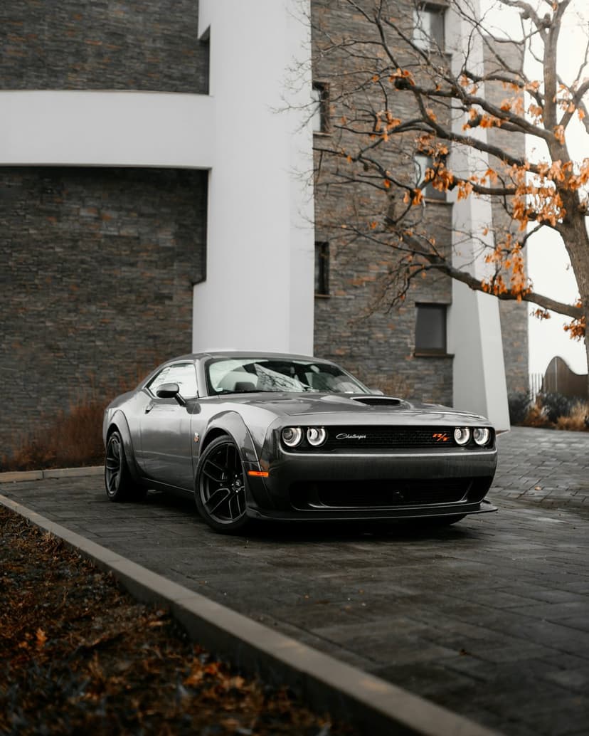 Grey Dodge Challenger Widebody parked on a concrete driveway near a modern brick building and autumn tree.
