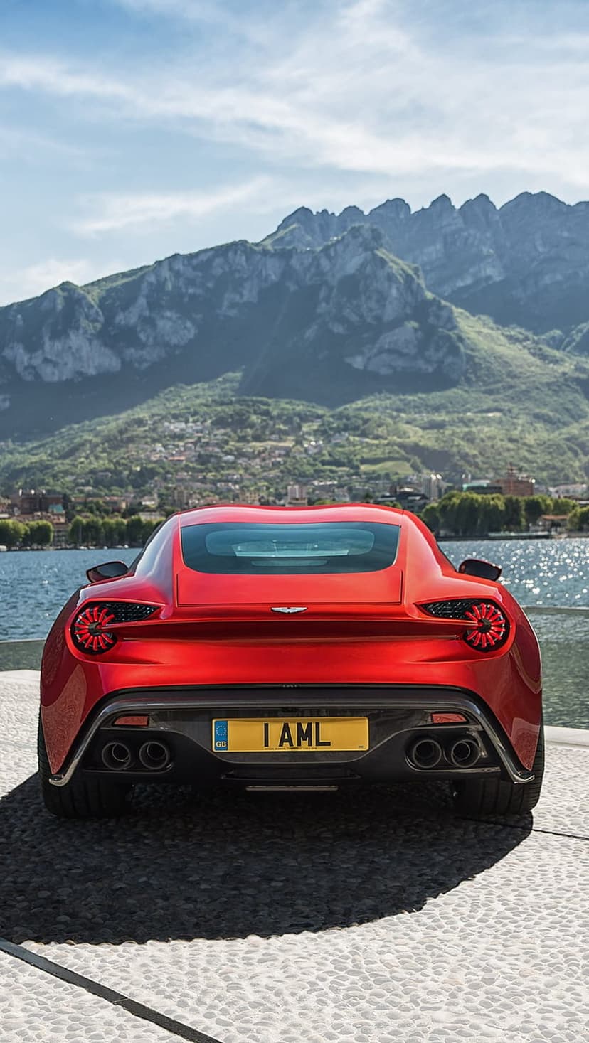 Red Aston Martin rear view parked near a mountain lake under a blue sky with a distant village in view.