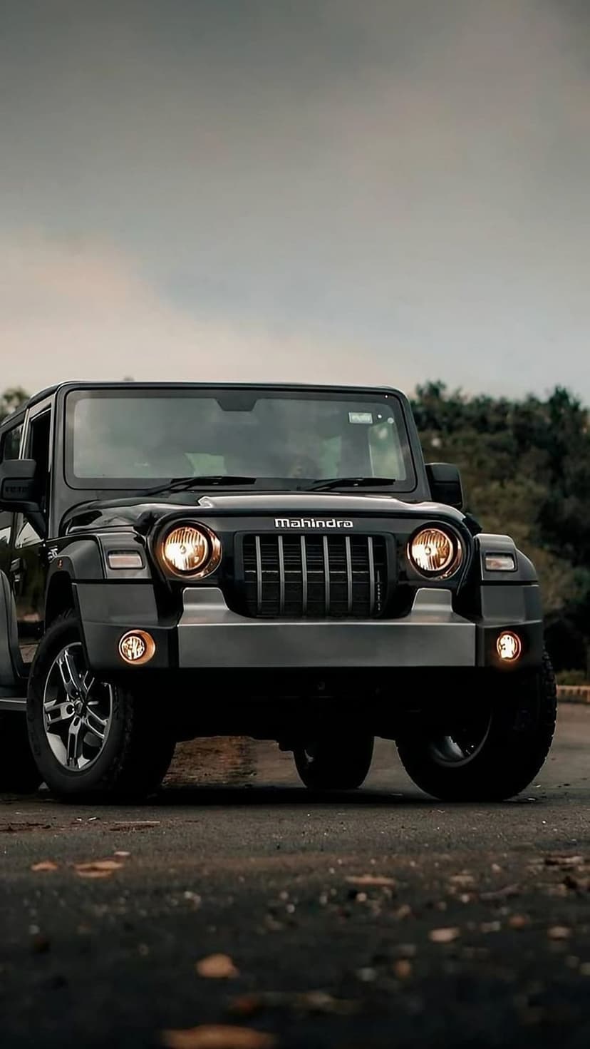 Black Mahindra Thar SUV with round headlights and silver bumper parked on an asphalt road with green trees behind.