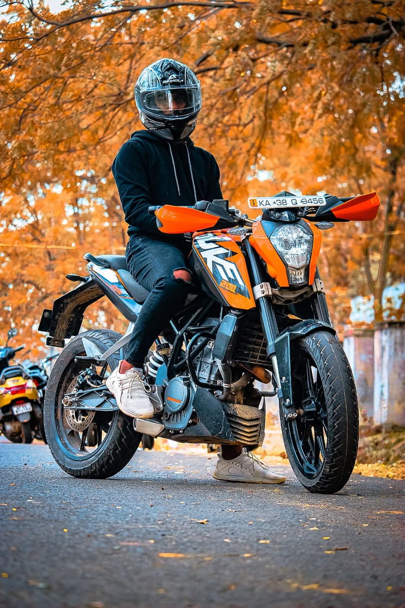 Biker in a black hoodie on an orange KTM motorcycle parked within a forest of golden autumn deciduous trees.