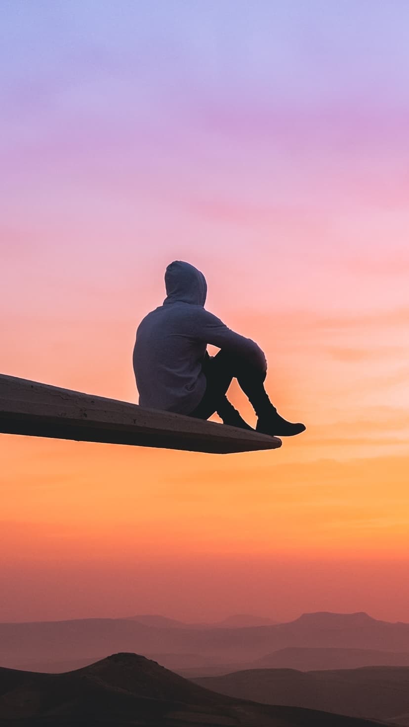 Silhouetted person sits on a wooden beam overlooking rolling hills under a vibrant purple and orange twilight sky.