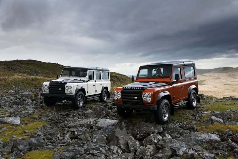 White and orange Land Rover Defenders parked on a rocky mountain slope under a dramatic cloudy sky.