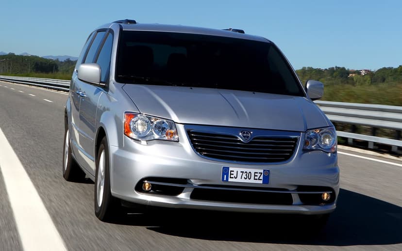 Silver Lancia Voyager driving on an asphalt highway under a clear blue sky with green hills in the distance.