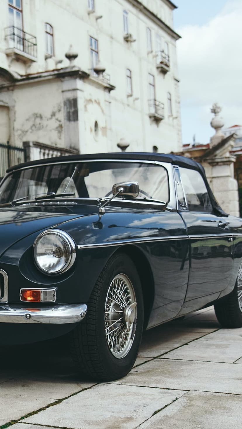 Dark blue vintage convertible parked on stone pavers in front of a grand manor with ornate windows and stonework.