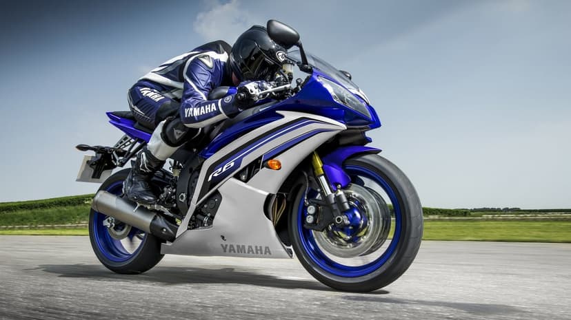 Blue Yamaha R6 motorcycle leaning into a turn with a rider in blue racing leathers on a paved track under sunlight.