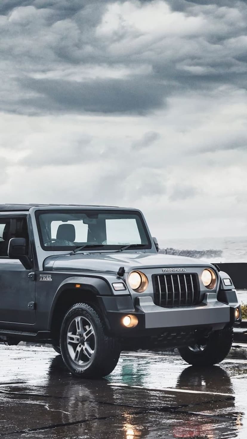 Mahindra Thar SUV parked on a reflective wet road with glowing headlights under a dark, cloudy sky.
