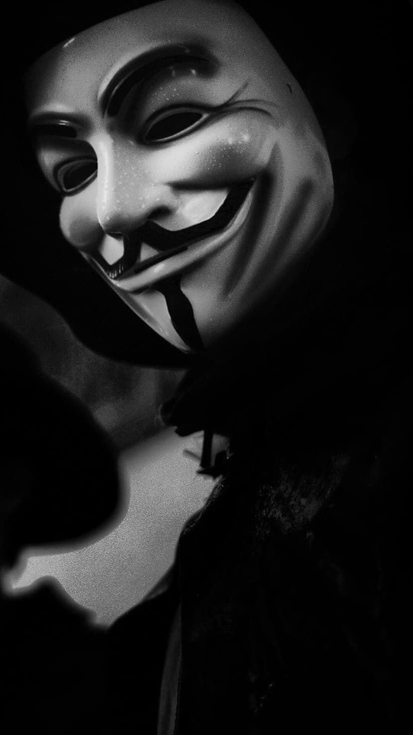 Guy Fawkes mask partially illuminated by harsh light against a pitch-black background in a vertical composition.