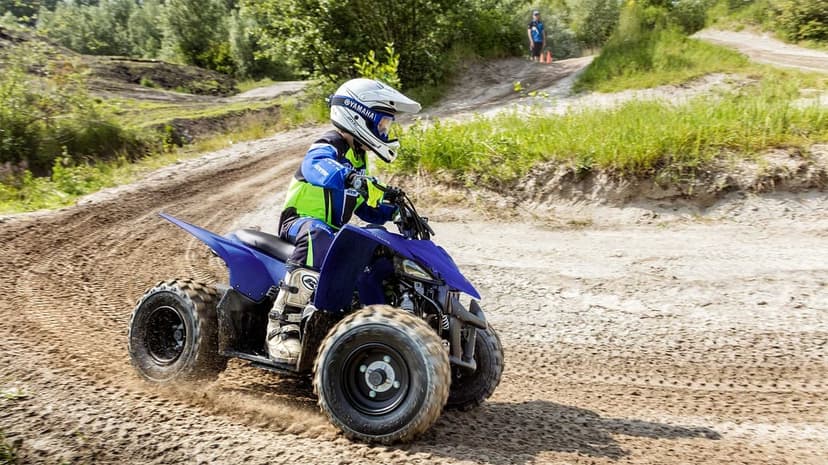 A young rider on a blue Yamaha ATV navigates a dusty dirt turn with a forest background in bright daylight.