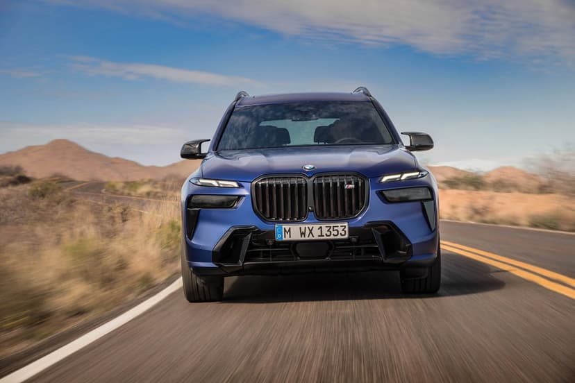 Front view of a blue BMW X7 M60i SUV driving on a desert road with motion blur and mountains background.